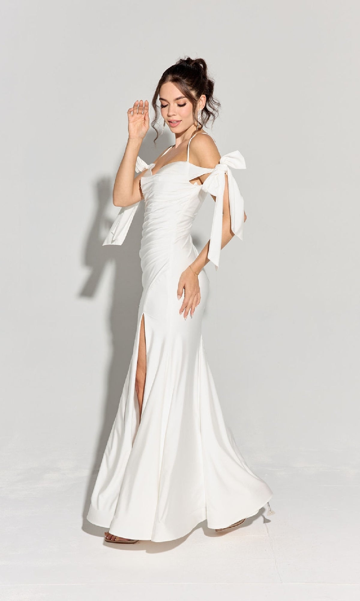 A woman stands against a plain background, showcasing the elegance of the affordable Shoulder-Ties Long Jersey Prom Dress: Eureka 9181, featuring bow details on the sleeves and a thigh-high slit.